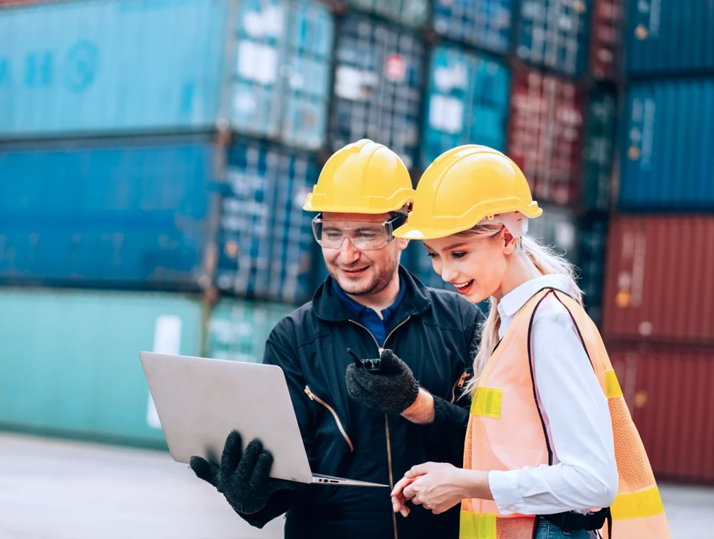 Two safety workers wearing helmets and vests smiling while looking at a laptop, standing in front of stacked shipping containers